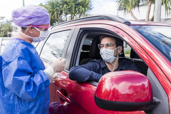 Man in red vehicle being tested for COVID-19 by a medical professional. Both are wearing masks.